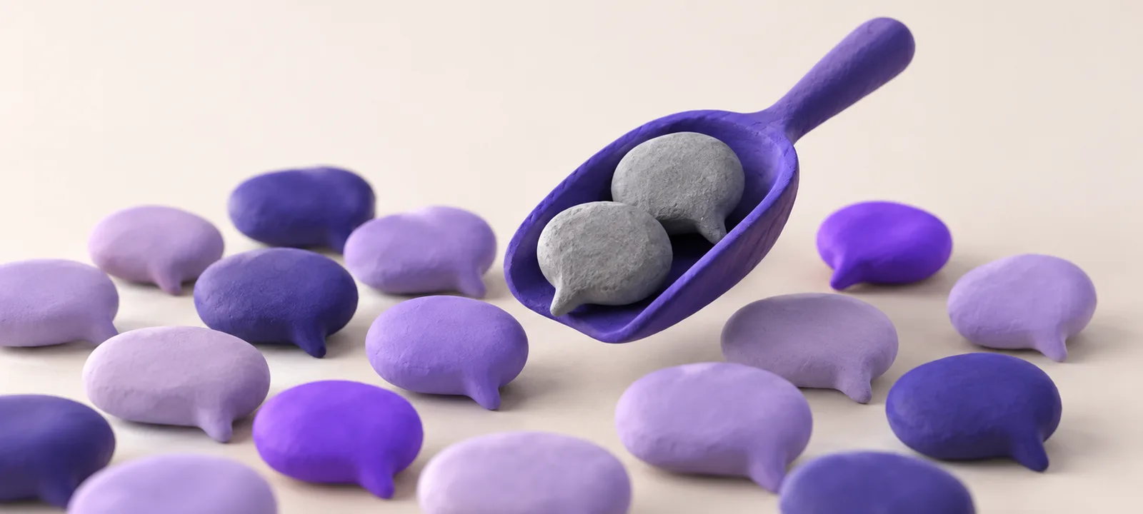 Small rounded clay speech-bubble shapes on a surface, mostly lavender and violet, while a clay scoop lifts two rough grey ones away